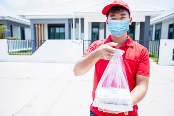 Asian delivery servicemen wearing a red uniform with a red cap and face mask handling food boxes in plastic bags to give to the customer in front of the house. Online shopping and Express delivery