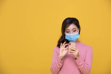 Koronavirüs krizi veya COVID-19 salgınına karşı maske takan Asyalı güzel, mutlu bir kadın portresi. Sarı arka planda cep telefonu ya da akıllı telefon kullanıyor.
