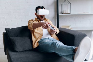 Young smiling man in VR headset playing video game with joystick, lying on the couch