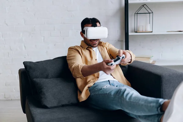 Man playing mobile game app in virtual reality glasses, holding joystick and lying on the couch — Stockfoto