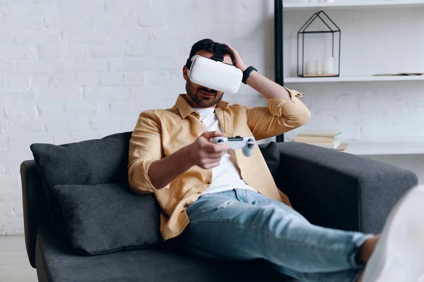 Young smiling man in VR headset playing video game with joystick, lying on the couch — Stockfoto