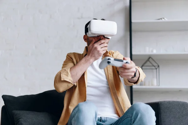 Young emotional man in VR glasses playing video game with joystick, cover his mouth with hand — Stockfoto