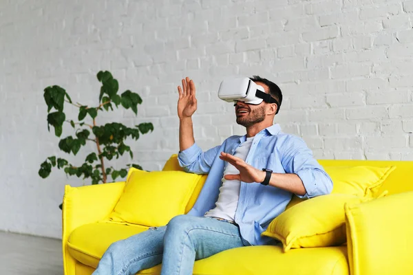 Front view of young smiling man using virtual reality glasses and showing his hand up — Stockfoto