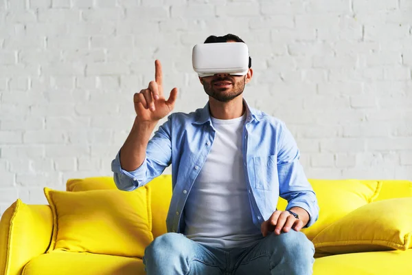 Front view of young smiling man using virtual reality glasses and showing his hand up — Stockfoto