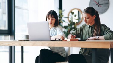Two young woman working in modern office using laptop and smart phone, making notes
