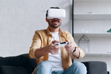 Young smiling man in VR glasses playing video game with joystick, siting on the couch
