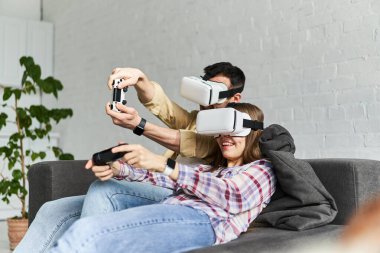 VR gözlüklü genç bir çift oyun oynuyorlar, kanepede oturuyorlar.