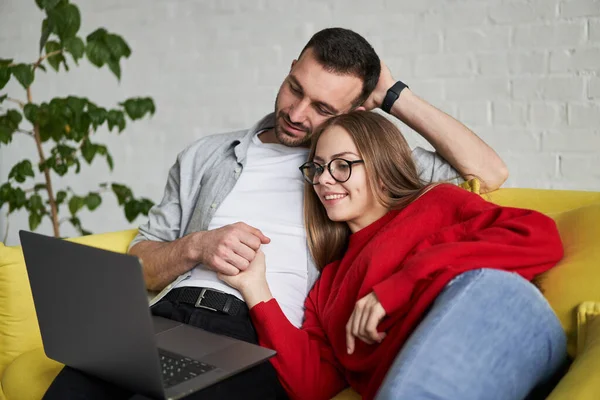 Close-up uitzicht van jong schattig paar op zoek naar laptop scherm gelegen op de gele bank — Stockfoto