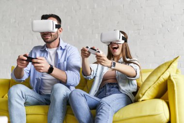 VR gözlüklü genç bir çift oyun oynuyorlar, kanepede oturuyorlar.