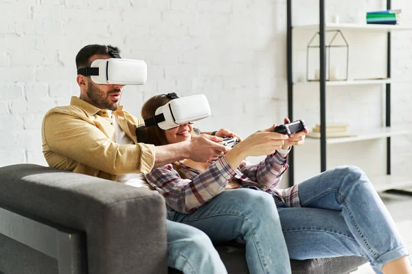 Young smiling couple in VR glasses playing video game with joystick, siting on the couch