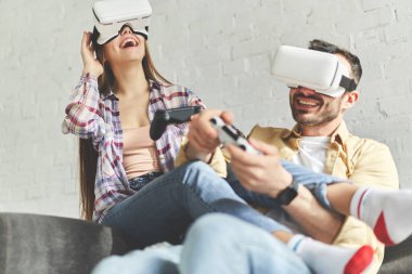 VR gözlüklü genç bir çift kanepede video oyunu oynuyor.