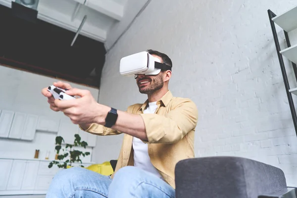 Zijaanzicht van de jonge lachende man in VR bril spelen video game met joystick, zittend op de bank — Stockfoto