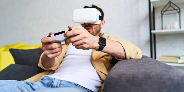 Close-up zicht van de jonge lachende man in VR bril spelen video game holding joystick, zittend op de bank — Stockfoto