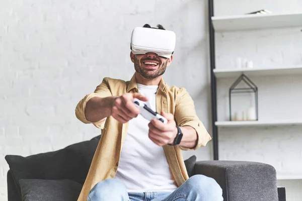 Young smiling man in VR glasses playing video game with joystick, siting on the couch — Stockfoto