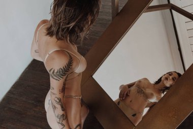 Aynanın yanında iç çamaşırlı, dövmeli seksi bir kadın.