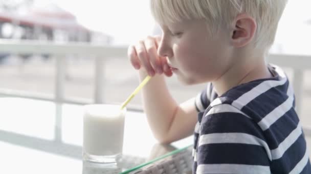 Aranyos szőke fiú tejturmixot iszik egy kávézóban egy forró nyári napon. — Stock videók