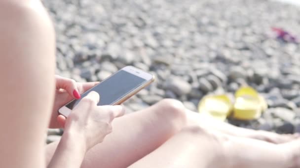 Μια γυναίκα διαβάζει με ένα κινητό τηλέφωνο σε μια παραλία με βότσαλα δίπλα στη θάλασσα — Αρχείο Βίντεο