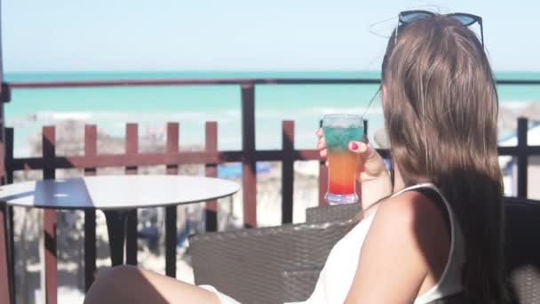 Una mujer bebe un fuerte cóctel alcohólico en una mesa de café junto al océano. — Vídeos de Stock