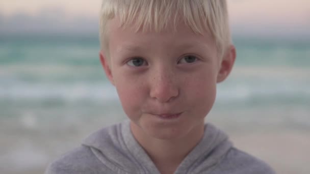 Κοντινό πλάνο ενός όμορφου ξανθού αγοριού στο φόντο μιας ξεπλυμένης θάλασσας — Αρχείο Βίντεο