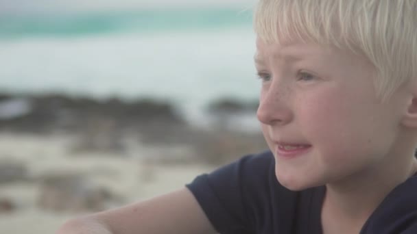 Vacker blond pojke sitter på stranden vid havet. — Stockvideo