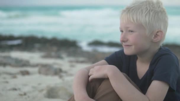 Un chico rubio se sienta en la playa junto al océano — Vídeos de Stock