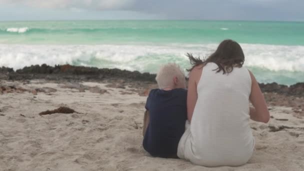 Mom and son sit on the beach by the ocean with their backs to the camera — Stock Video