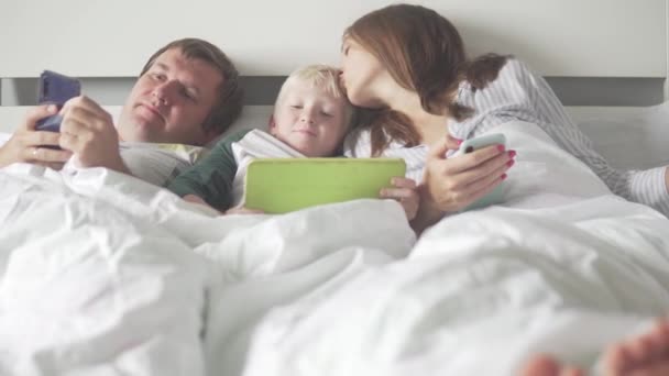 La familia se encuentra en una cama con una tableta y teléfonos en casa — Vídeo de stock