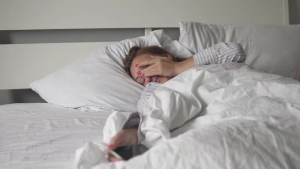 Woman wakes up in the morning in bed and watches time on a mobile phone — 비디오