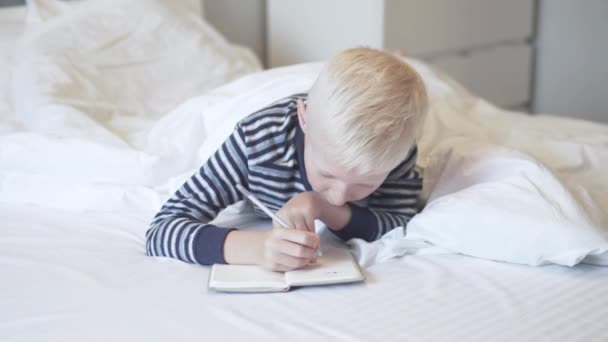 O rapaz loiro deita-se na cama com um caderno e uma caneta de manhã — Vídeo de Stock