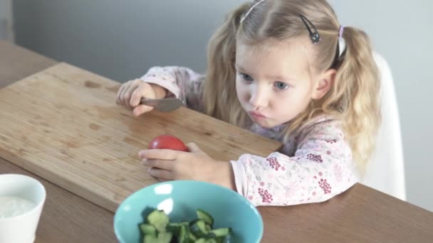 Ένα όμορφο κοριτσάκι κόβει μια ντομάτα για μια σαλάτα λαχανικών. — Αρχείο Βίντεο