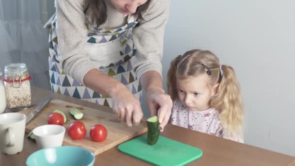 Мама вчить свою маленьку дочку зрізати огірки для салату. — стокове відео