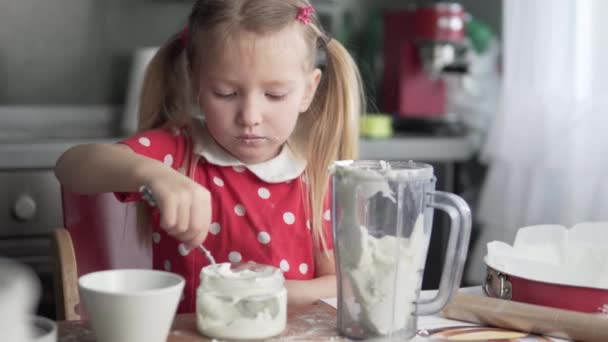 A cute little girl licks a spoon with cake cream — Stock Video