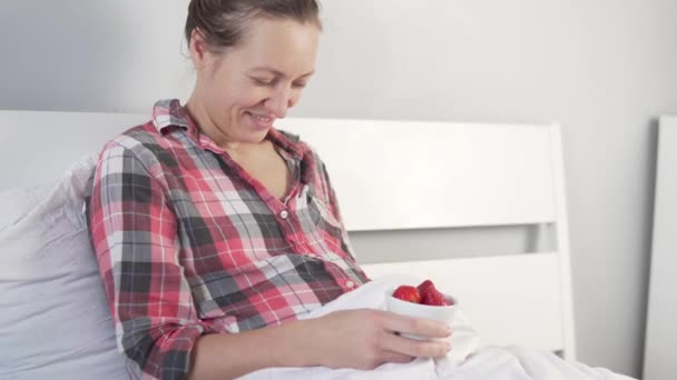 Беременная женщина ест клубнику в постели — стоковое видео