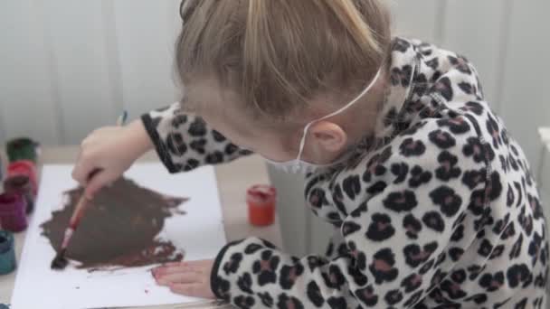 En liten flicka i en medicinsk mask på ansiktet målar hemma. — Stockvideo