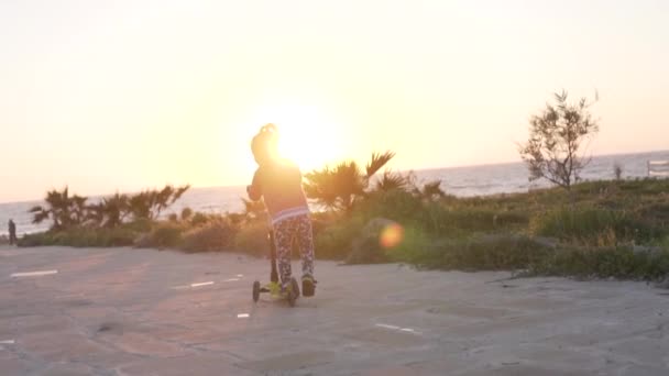 En liten flicka åker skoter längs havet på kvällen — Stockvideo