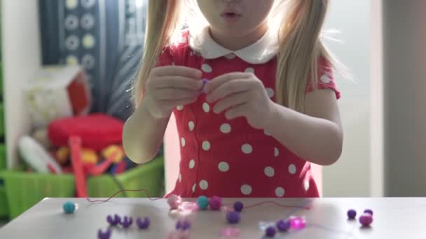 Una bambina fa un braccialetto di perline a casa a tavola — Video Stock