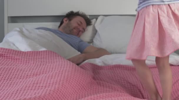 A little girl jumps on a bed next to her sleeping fathe — Stock Video