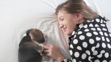 Sevimli bir köpek yavrusu olan bir kadın yatağa uzanıyor ve köpeği okşuyor.