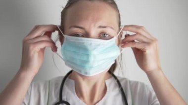 Yorgun bir kadın doktor yüzüne tek kullanımlık tıbbi bir maske takıyor.