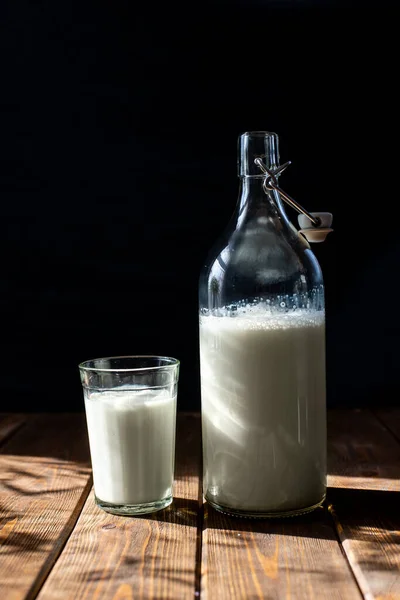 Μπουκάλι και ποτήρι γάλα σε καφέ ξύλινο τραπέζι. σκοτεινές ακτίνες ήλιου υποβάθρου. κτηνοτροφικά προϊόντα υγιεινή διατροφή — Φωτογραφία Αρχείου