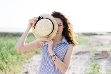 Vahşi bir sahilde duran bir kadın. Çiller, siyah bukleler, mavi bir elbise, hasır şapka.