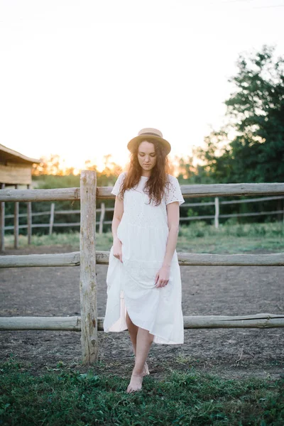 美しいブルネットの女の子の白いドレスでわらの帽子馬のパドックの近くに立って — ストック写真