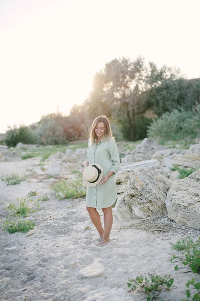 ドレスの女性は、野生のビーチ、石の上に立っている。ブロンド、藁帽子 — ストック写真