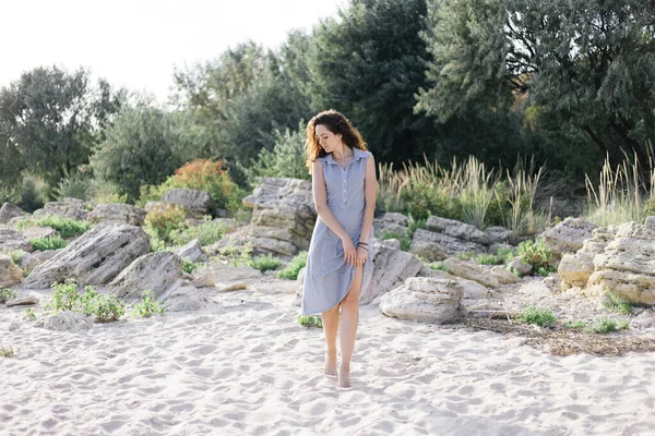 女性は野生のビーチ、石の上に立っている。そばかす暗いカールの髪青いドレス — ストック写真