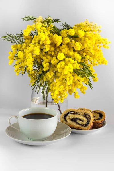 Vase with mimosa branches, cup of coffee and poppy seed strudel