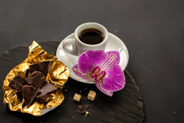 Bir fincan sade kahve, altın folyoda çikolata ve siyah arka planda siyah bir sayfada bir orkide çiçeği..