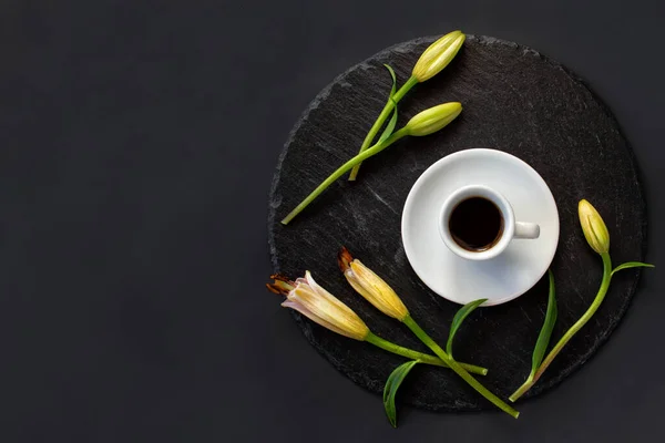 Cup of black coffee and fresh spring buds of lily flowers on a round slate on a black background. Romantic stylish breakfast. Date on restaurant. Still life flat lay. Top view. Space for text.