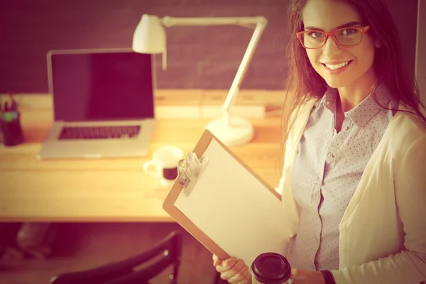 Giovane donna in piedi vicino alla scrivania con cartella del computer portatile e tazza di caffè — Foto Stock