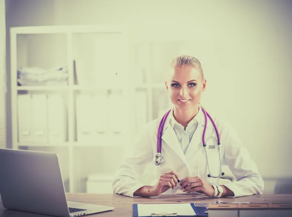 Bonito jovem sorridente médico feminino sentado na mesa e escrevendo. — Fotografia de Stock