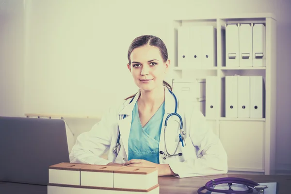 Mooie jonge glimlachende vrouwelijke arts zitten aan het bureau en schrijven. — Stockfoto